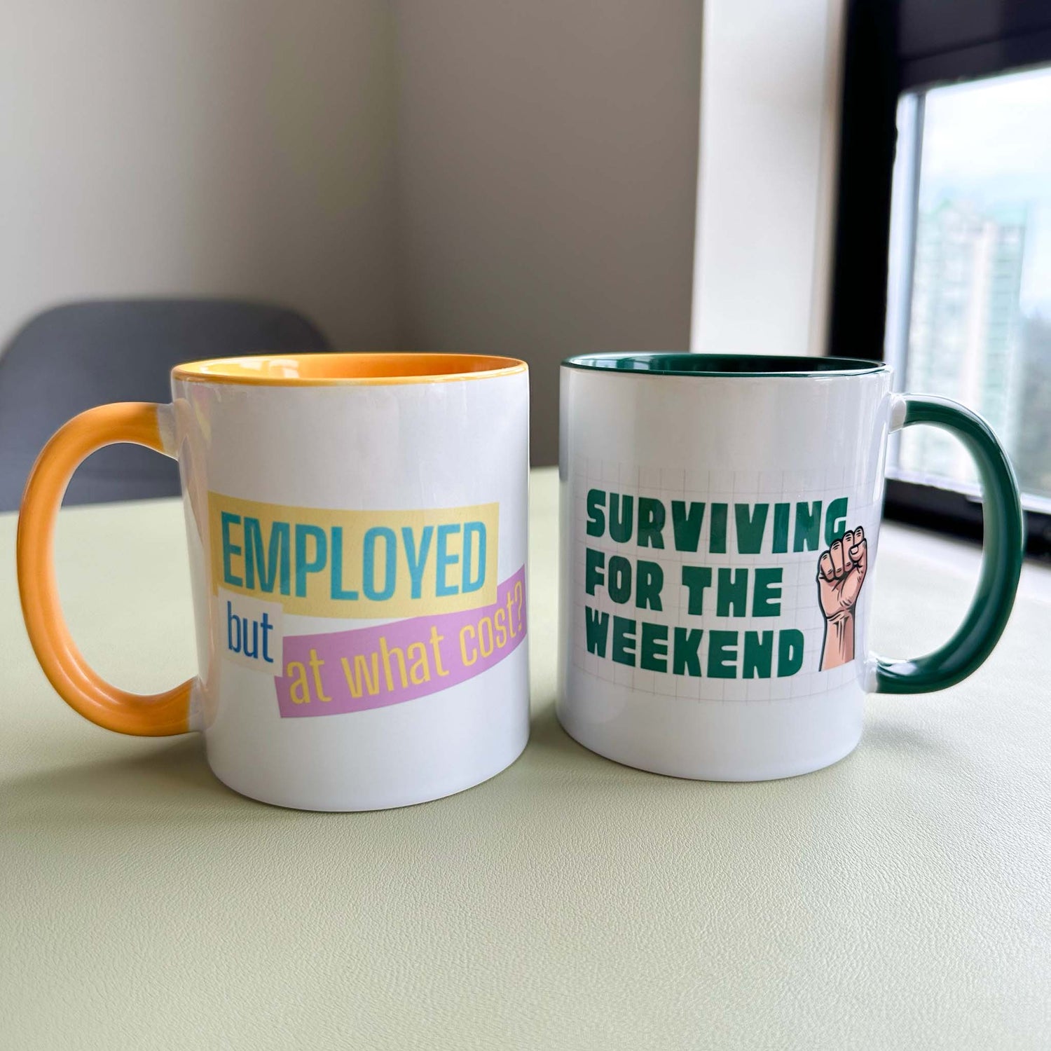 Two Deskbounders ceramic mugs side by side on a desk: 'Employed but at what cost?' with an orange interior and handle, and 'Surviving for the weekend' with a dark green interior and handle.