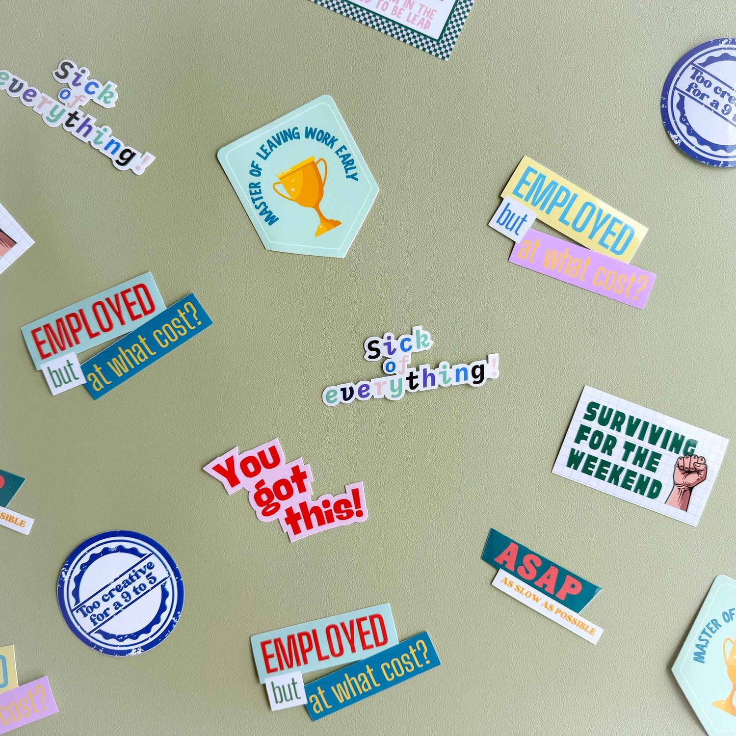 Close-up assortment of Deskbounders vinyl stickers scattered on a sage green surface, including 'Surviving for the weekend', 'Employed but at what cost?', 'You got this!', and 'ASAP as slow as possible'.