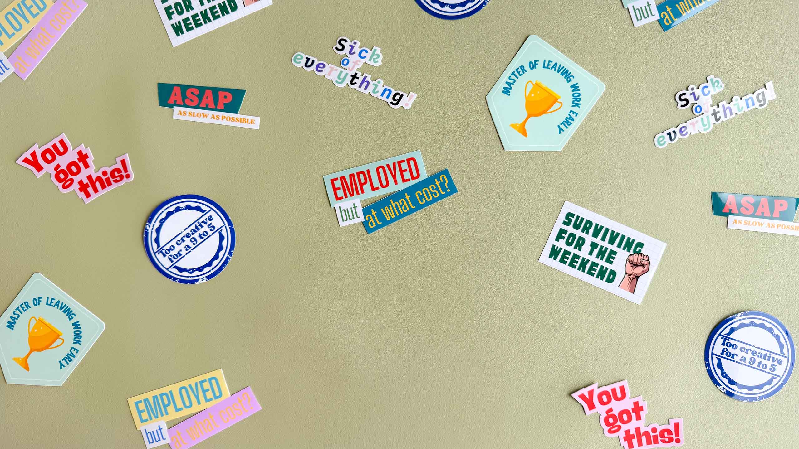 Assortment of Deskbounders vinyl stickers scattered on a sage green surface, including 'Surviving for the weekend', 'Employed but at what cost?', 'You got this!', 'Master of leaving work early', 'Sick of everything!', 'ASAP as slow as possible', and 'Too creative for a 9 to 5'.