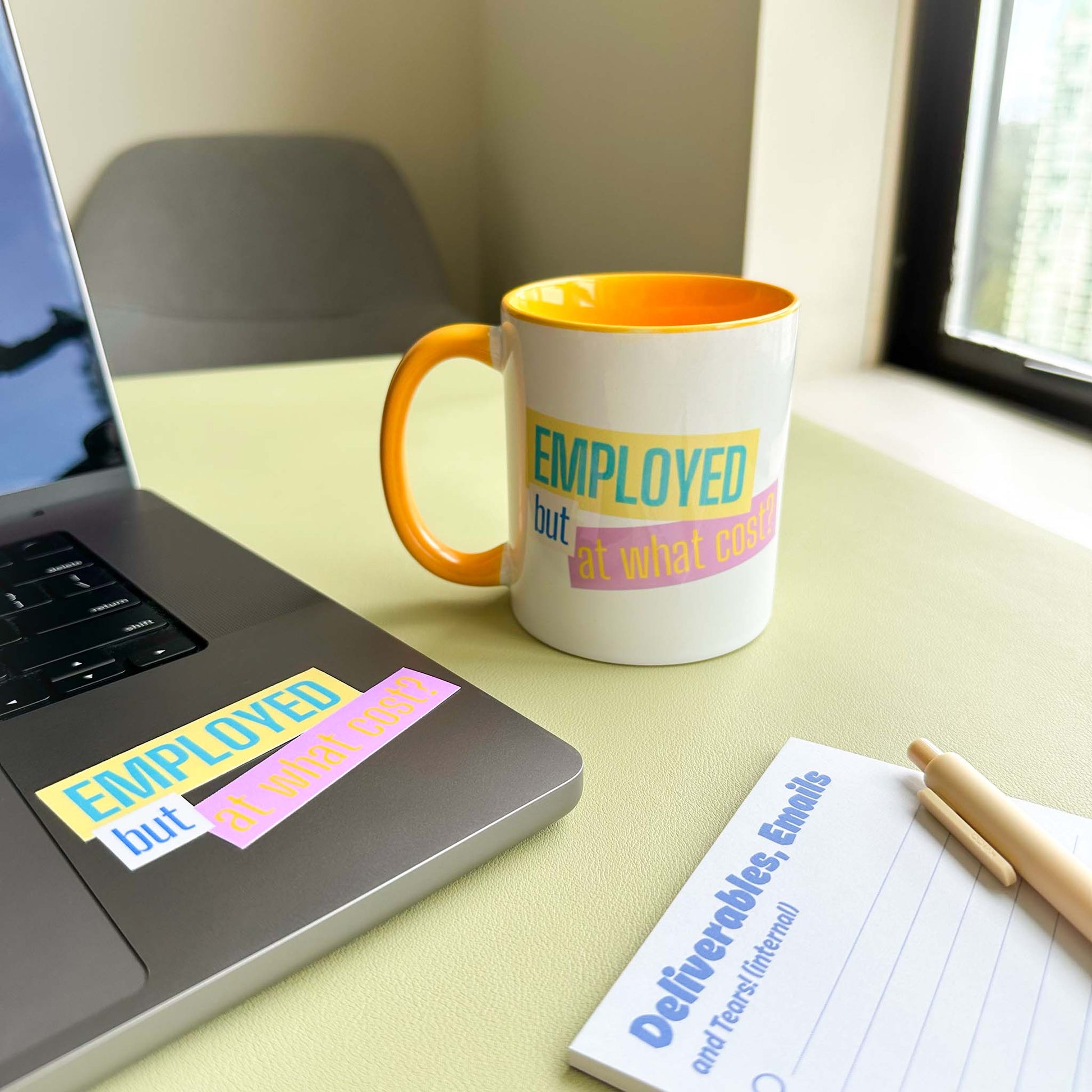 'Employed but at what cost?' ceramic mug on a desk beside a laptop with the matching sticker, and a 'Deliverables, Emails and Tears' notepad