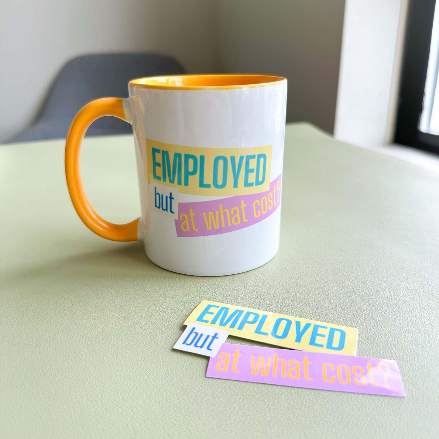 'Employed but at what cost?' ceramic mug paired with the matching vinyl sticker on a desk