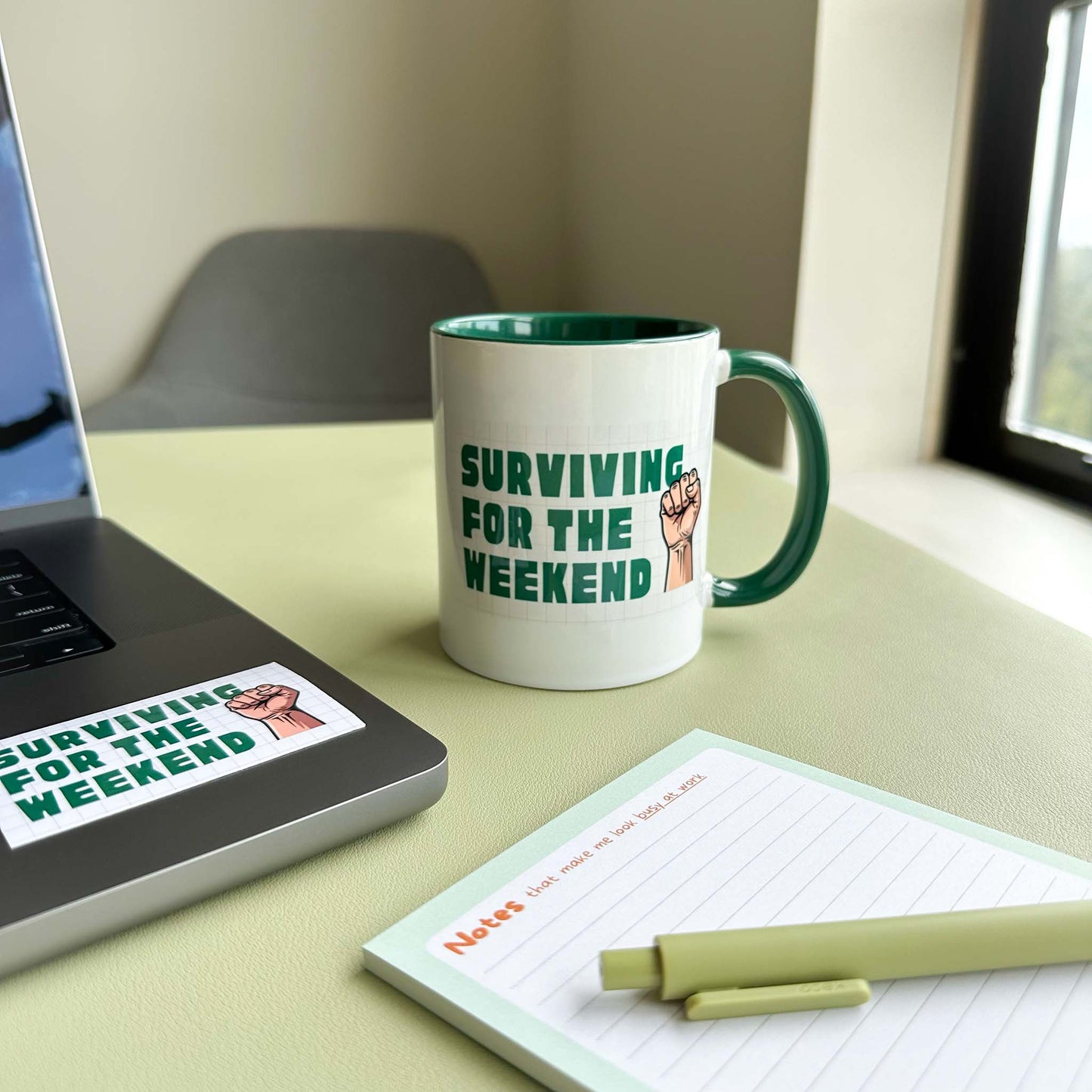 'Surviving for the weekend' ceramic mug on a desk beside a laptop with the matching sticker, a notepad, and a pen
