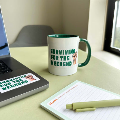 'Surviving for the weekend' ceramic mug on a desk beside a laptop with the matching sticker, a notepad, and a pen