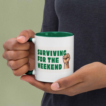 White ceramic mug with green interior reading 'Surviving for the weekend' in bold green text with a raised fist illustration